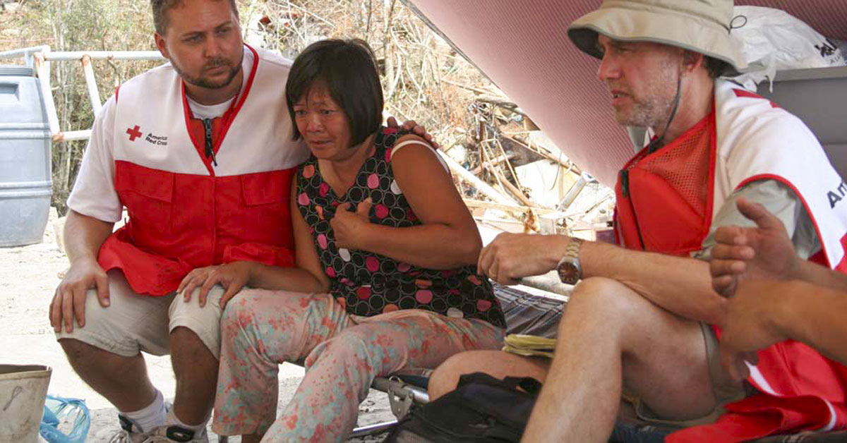 Distressed couple aided by a Red Cross helper in a covered shelter.