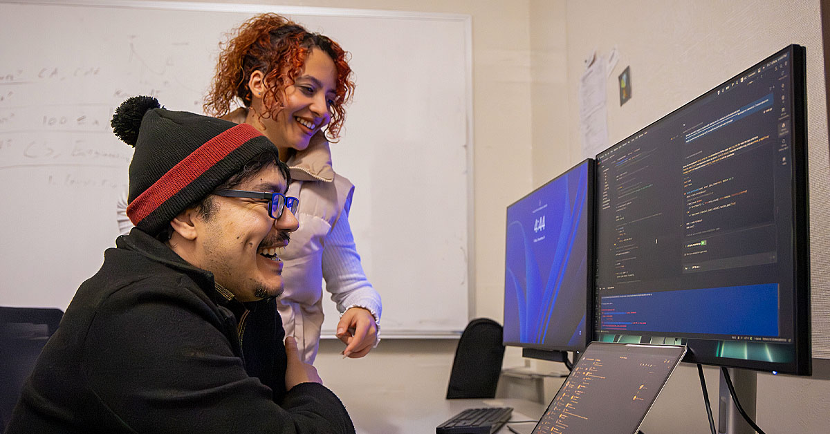 Two people smiling while reviewing code on dual monitors in an office, with a laptop open and a whiteboard in the background.